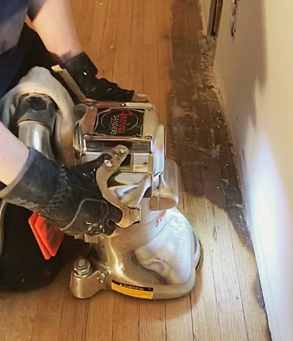 Close up of Alanna using the edge sander, a large handheld orbital sander, to refinish the flooring around the edged of the room.