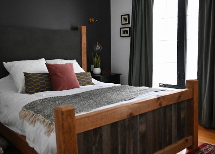 a moody bedroom with a dark accent wall and barnwood bed with white sheets and comfortable bedding. 